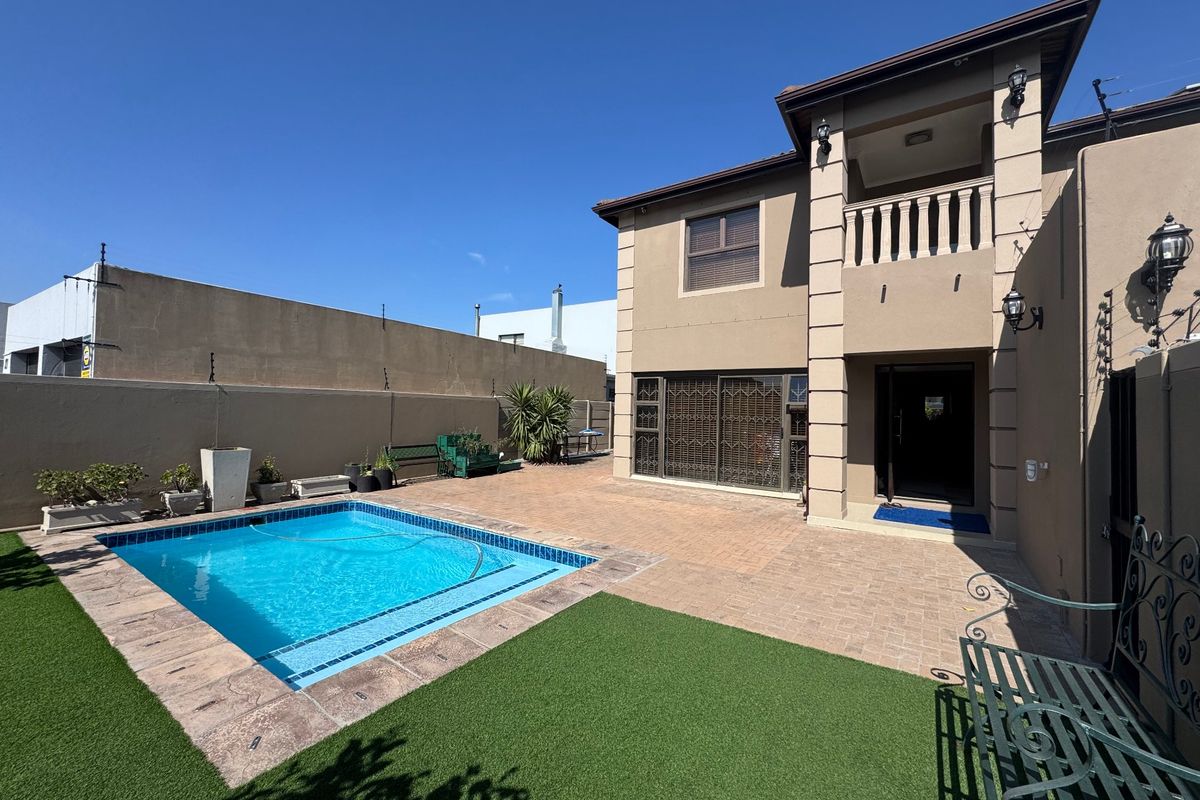 Rear exterior view featuring a swimming pool, paved patio area, artificial lawn, and a two-story building facade with a balcony.