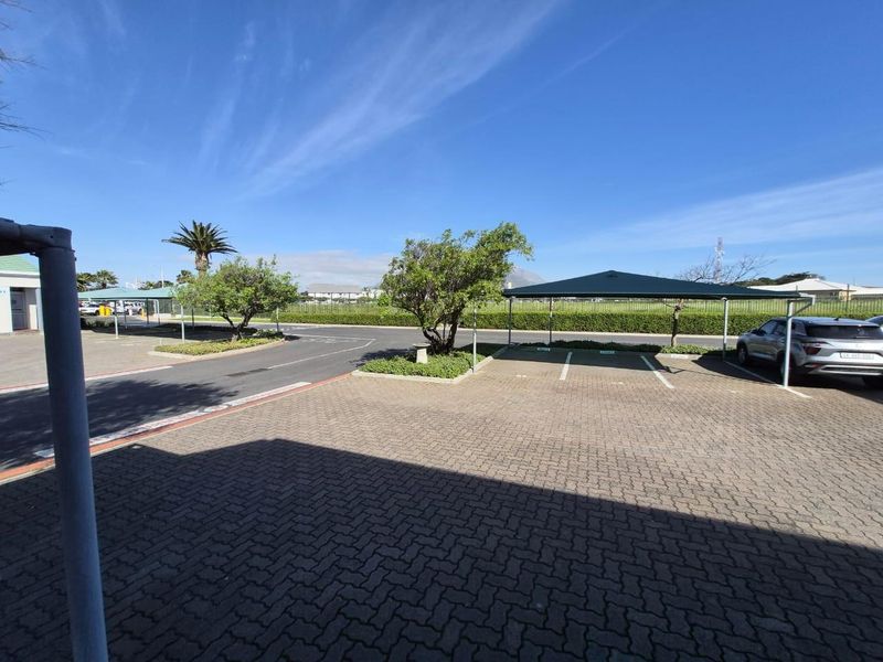 A well-maintained commercial parking area featuring paved bays, shade cloth carports, and landscaped garden beds under a clear blue sky.
