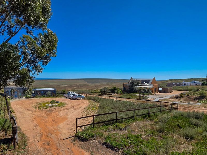 An aerial view of a rural property featuring a main dwelling with solar panels, outbuildings, and expansive surrounding land with gravel access.