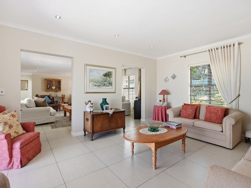 A spacious, light-filled living room featuring neutral tiled flooring, recessed downlighting, and large windows with venetian blinds, offering an open flow to adjacent areas.