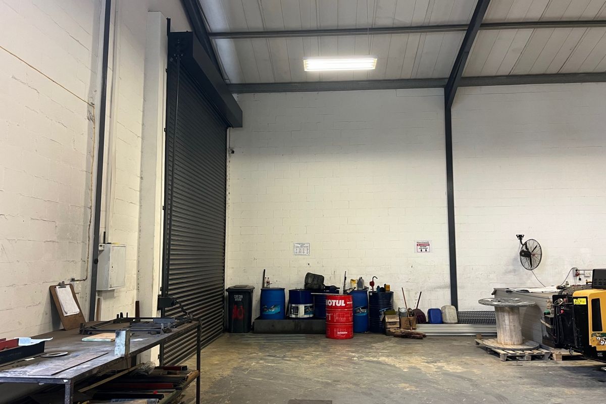 Spacious commercial utility or warehouse area featuring high ceilings, exposed steel rafters, concrete flooring, and a large roller shutter door for easy access.