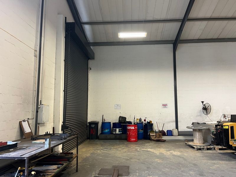 Spacious commercial utility or warehouse area featuring high ceilings, exposed steel rafters, concrete flooring, and a large roller shutter door for easy access.