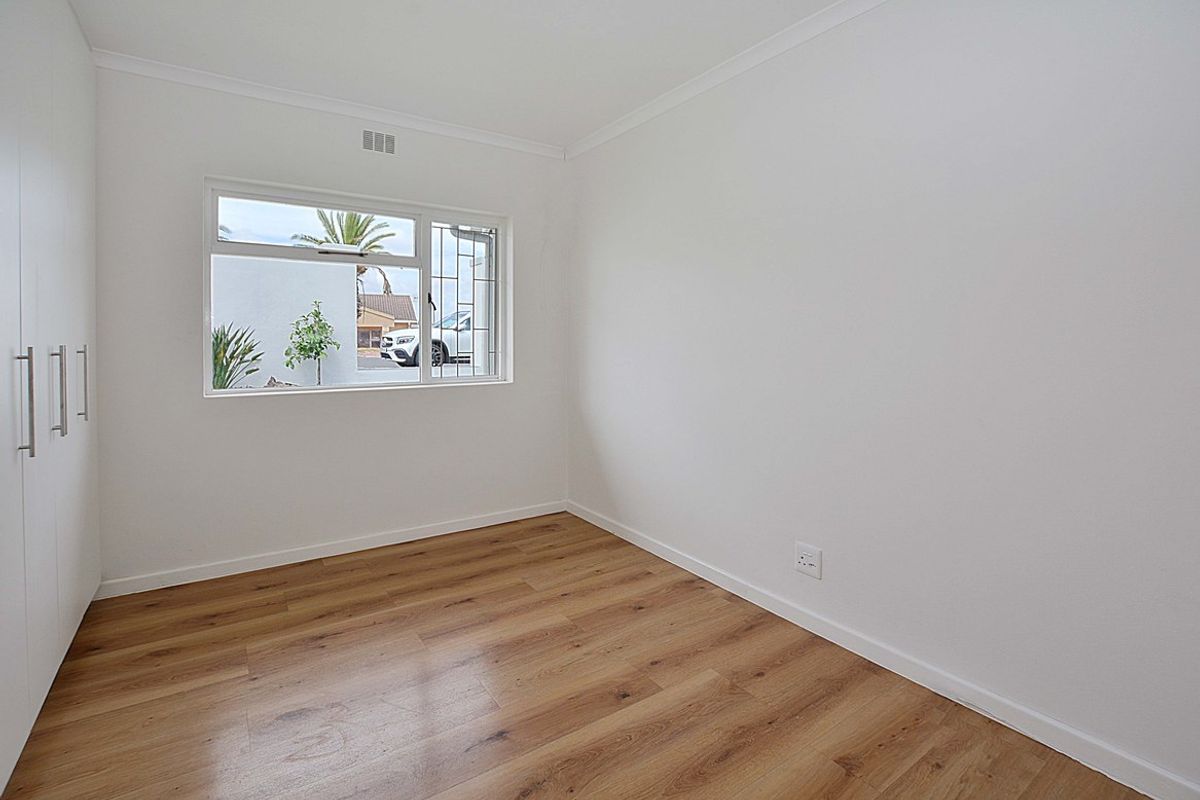 A bright, empty bedroom featuring modern wood-look laminate flooring, built-in cupboards, and a window with security bars.