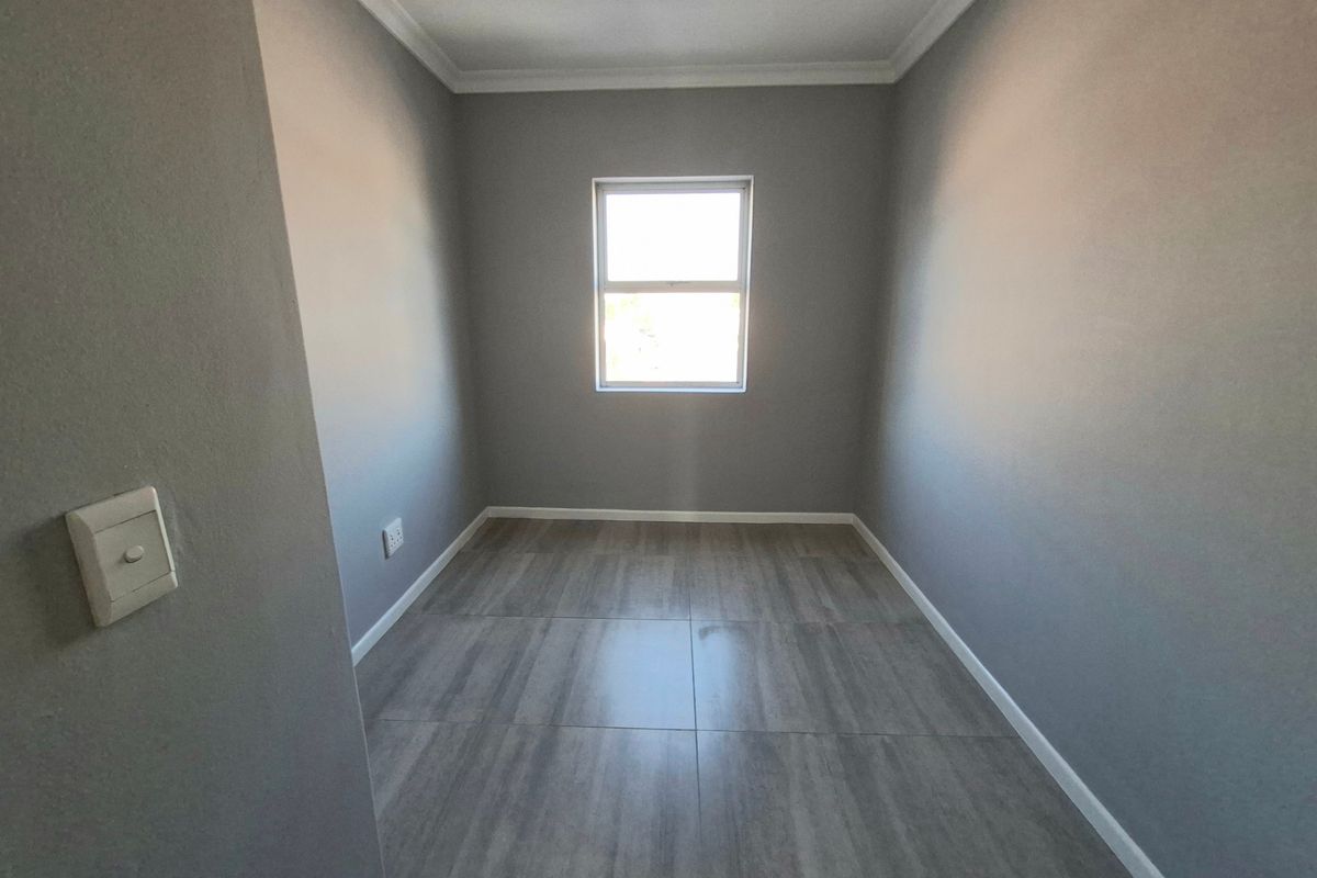 A clean, empty bedroom featuring neutral grey walls, modern wood-look floor tiles, and a central window providing natural light.