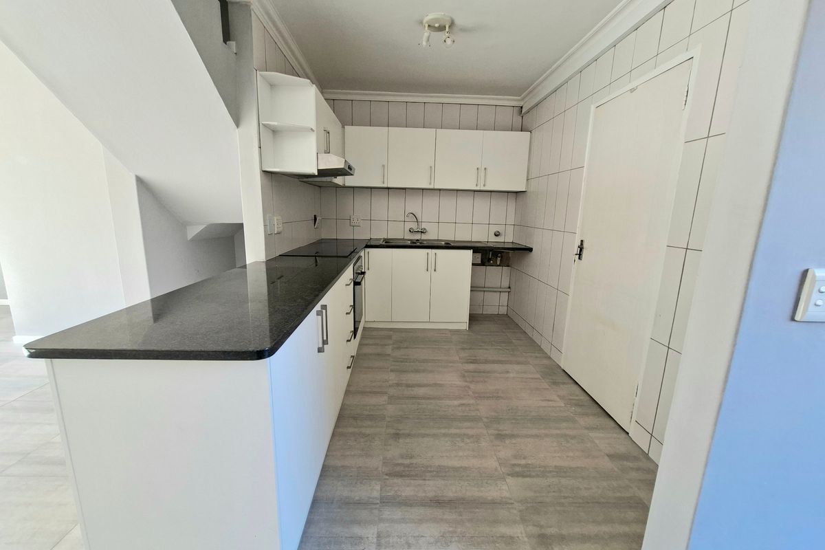A functional kitchen featuring white cabinetry, dark stone countertops, tiled walls, and grey floor tiles, situated adjacent to an internal staircase.