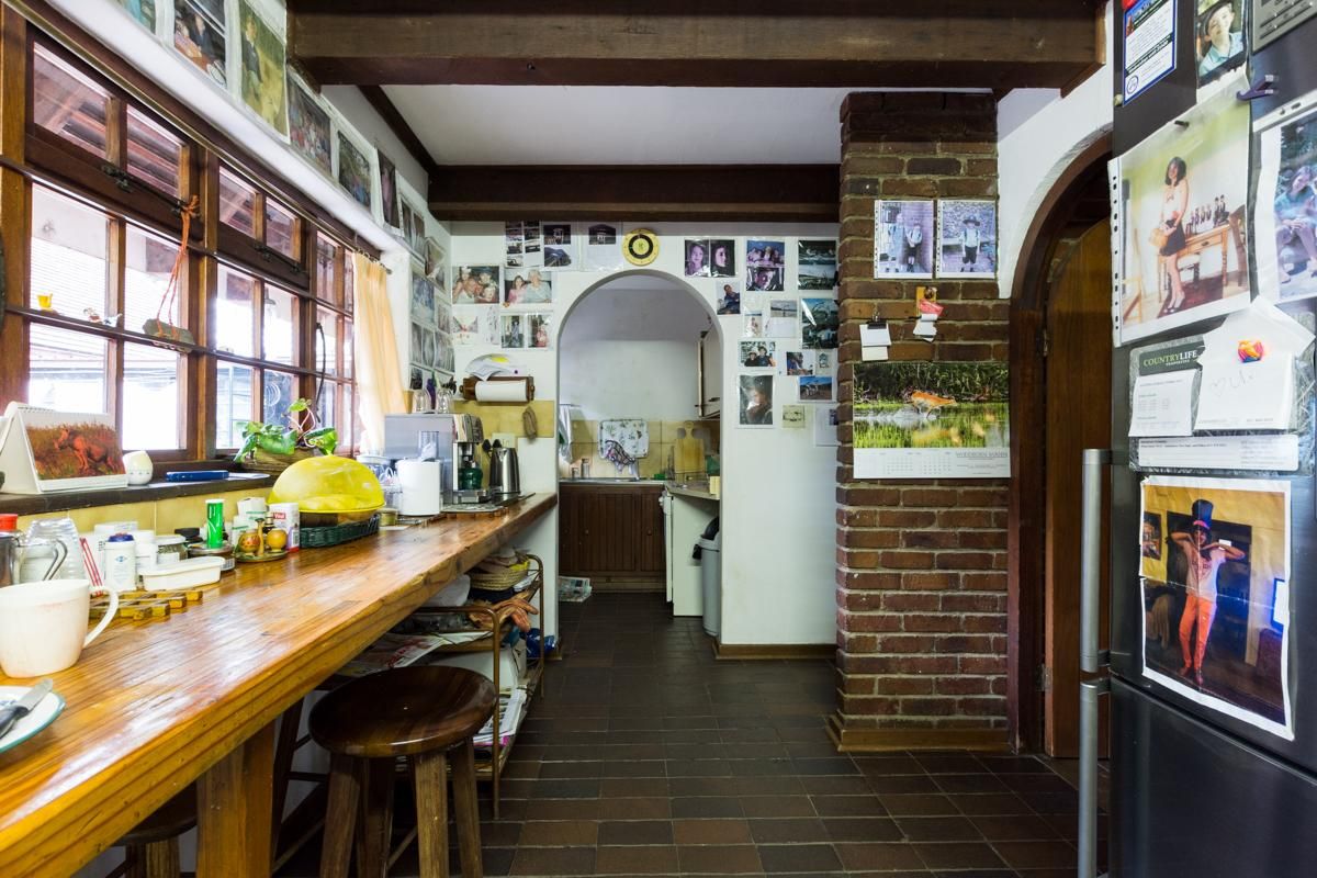 A rustic kitchen area featuring exposed brickwork, timber ceiling beams, a long wooden breakfast bar, and tiled flooring leading to a secondary prep space.