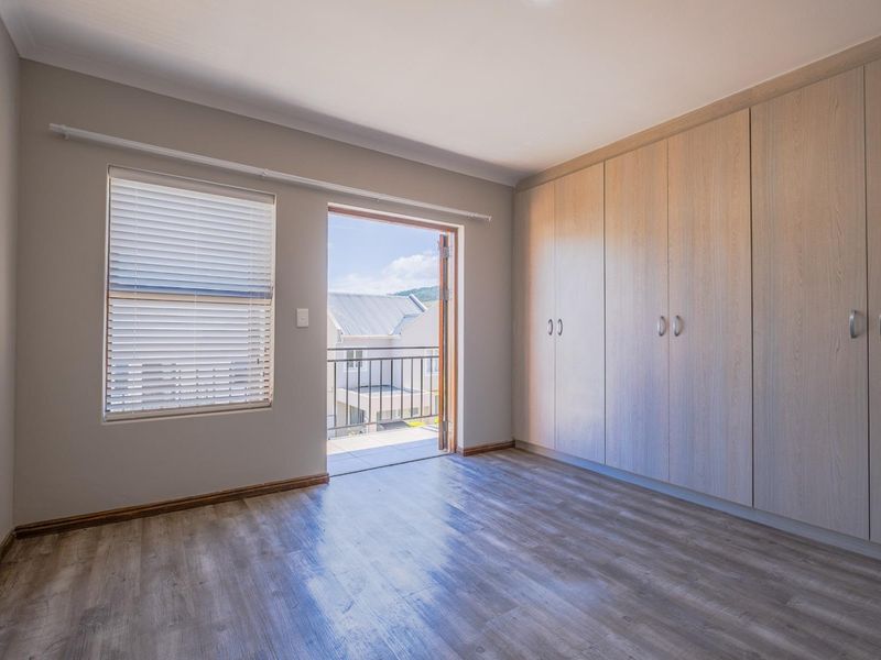 A bright, empty bedroom featuring light wood-tone built-in cupboards, modern laminate flooring, and direct access to a private balcony.