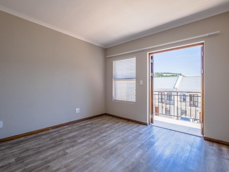 A bright, empty bedroom featuring modern laminate flooring, neutral walls, and double doors opening onto a private balcony.
