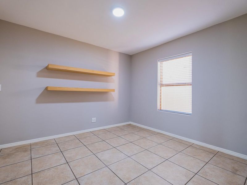 A clean, empty bedroom featuring neutral grey walls, tiled flooring, built-in floating shelves, and a window with fitted blinds.