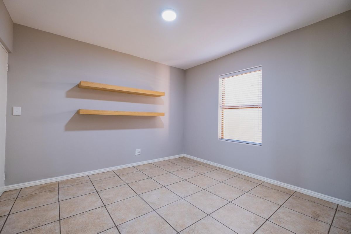 A clean, empty bedroom featuring neutral grey walls, tiled flooring, built-in floating shelves, and a window with fitted blinds.