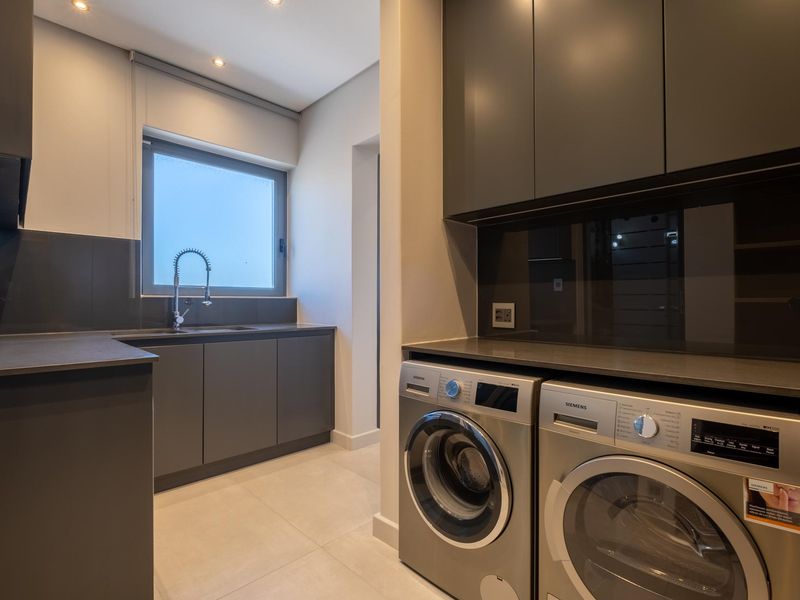 Modern laundry room featuring sleek dark cabinetry, stone countertops, a stainless steel sink with gooseneck faucet, and integrated space for laundry appliances.