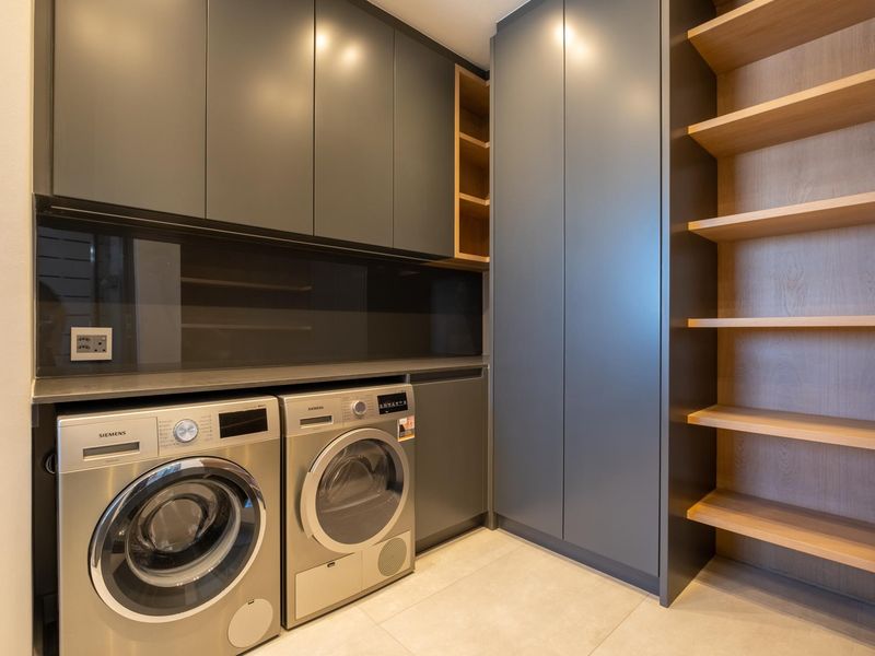 Modern laundry room featuring sleek dark grey cabinetry, integrated shelving, and space for side-by-side laundry appliances with a clean tiled floor.