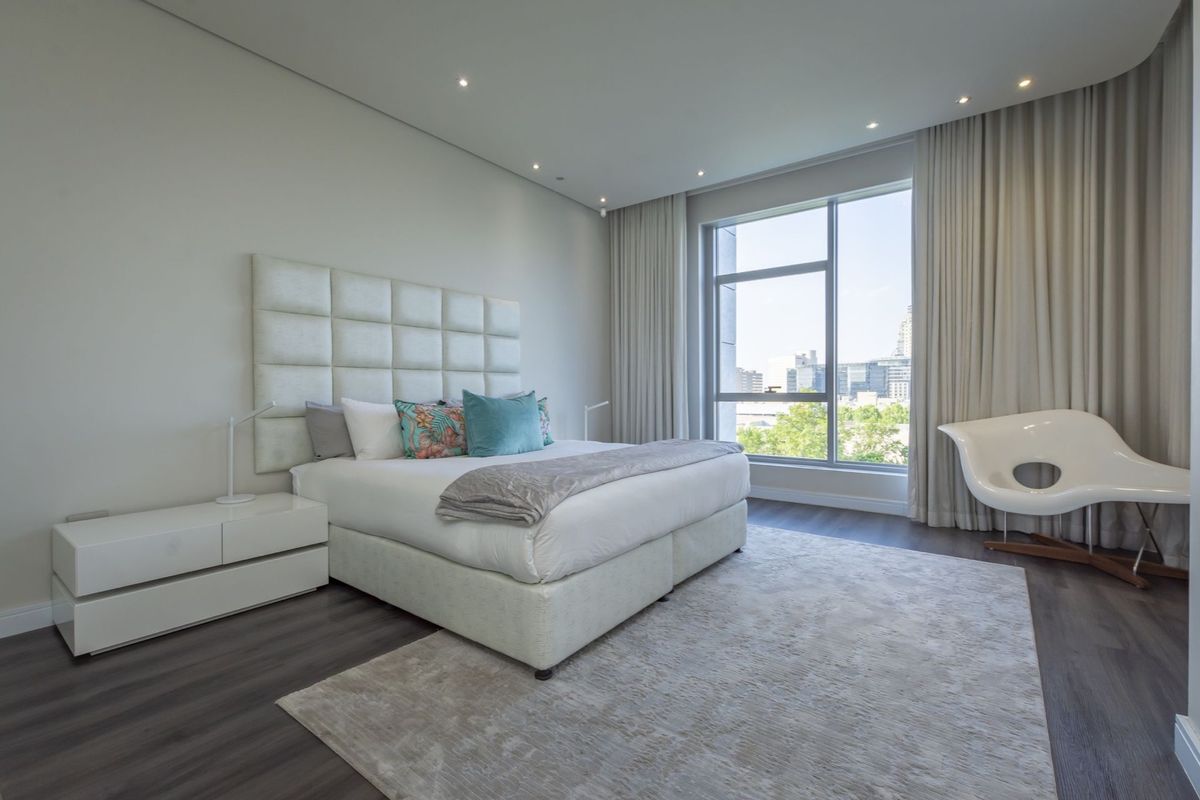 A modern, well-lit bedroom featuring dark wood-look flooring, recessed ceiling lighting, and large floor-to-ceiling windows with city views.