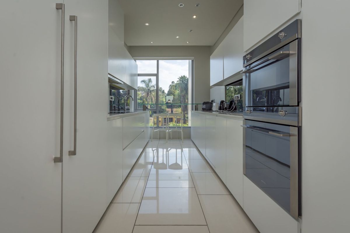 Modern galley-style kitchen featuring sleek white cabinetry, integrated double ovens, and polished floor tiles with a large window providing natural light.