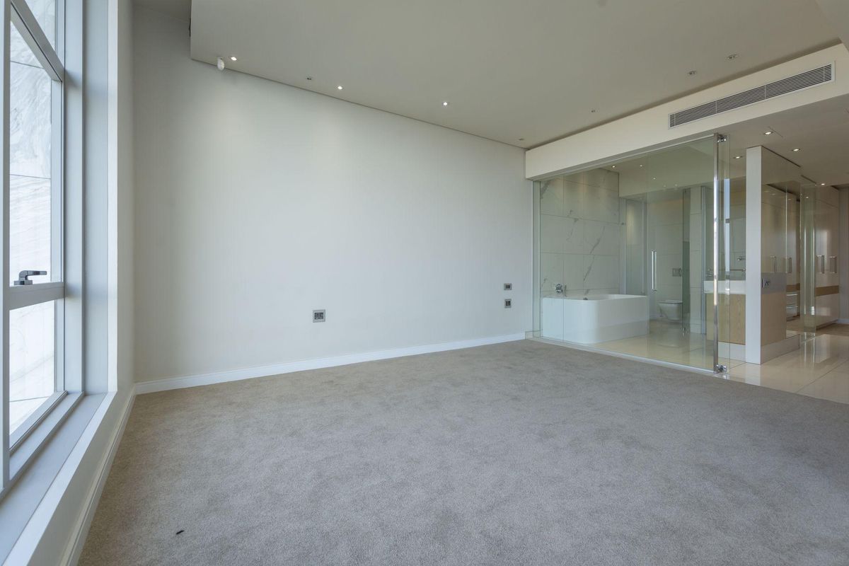A spacious, empty bedroom featuring neutral carpeting, recessed ceiling lighting, and an integrated glass-enclosed en-suite bathroom with modern finishes.