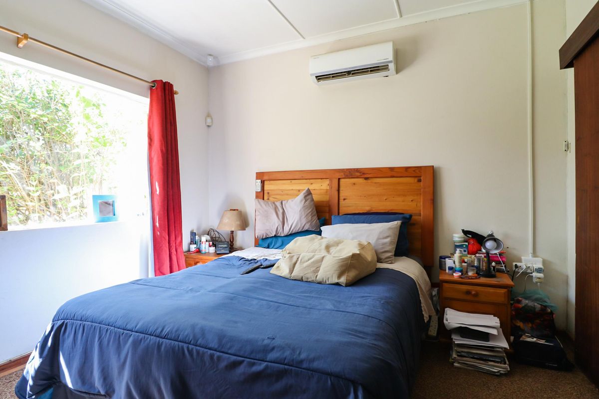 A functional bedroom featuring a wall-mounted air conditioning unit, large window for natural light, and carpeted flooring.