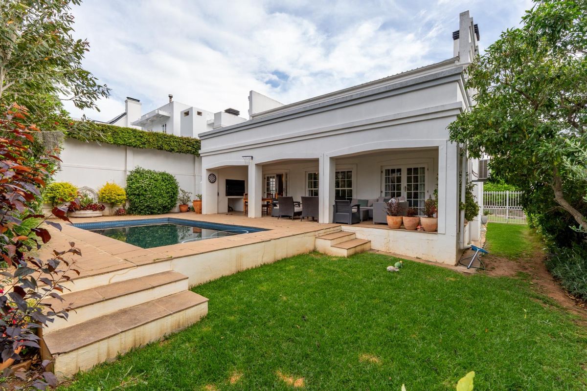 Rear exterior view featuring a covered patio, built-in braai area, and a swimming pool set within a landscaped garden.