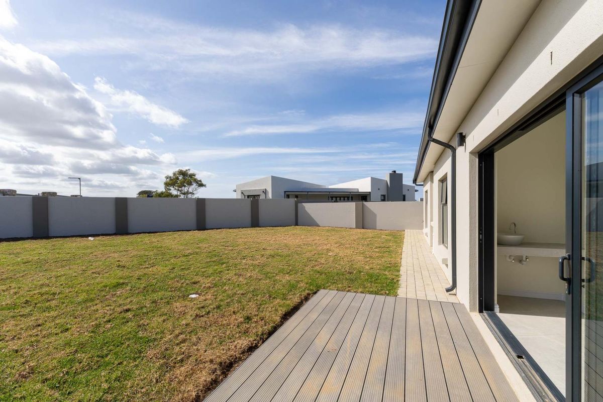 A view of the rear garden featuring a wooden deck, paved walkway, and perimeter boundary wall, accessible via sliding glass doors.