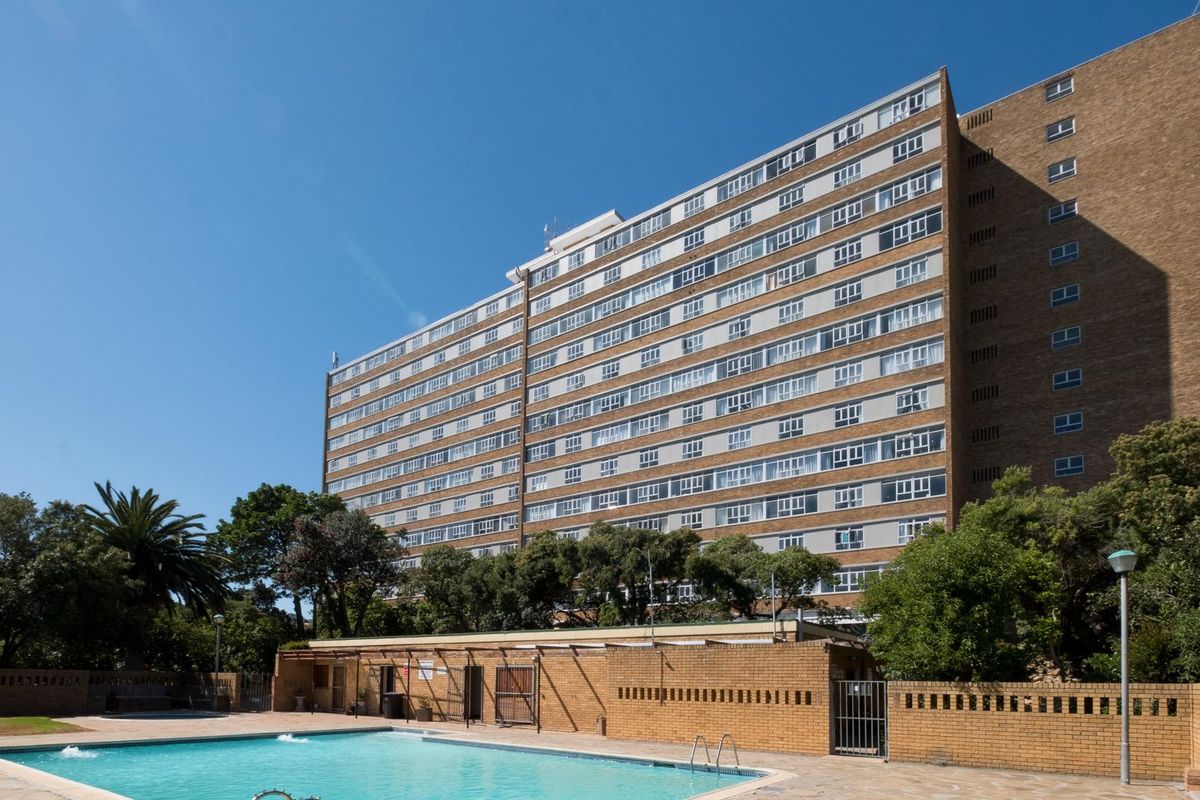 A well-maintained communal swimming pool area set against a large multi-story apartment building with established trees and brick perimeter walls.
