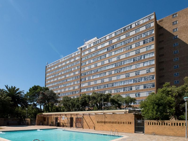 A well-maintained communal swimming pool area set against a large multi-story apartment building with established trees and brick perimeter walls.
