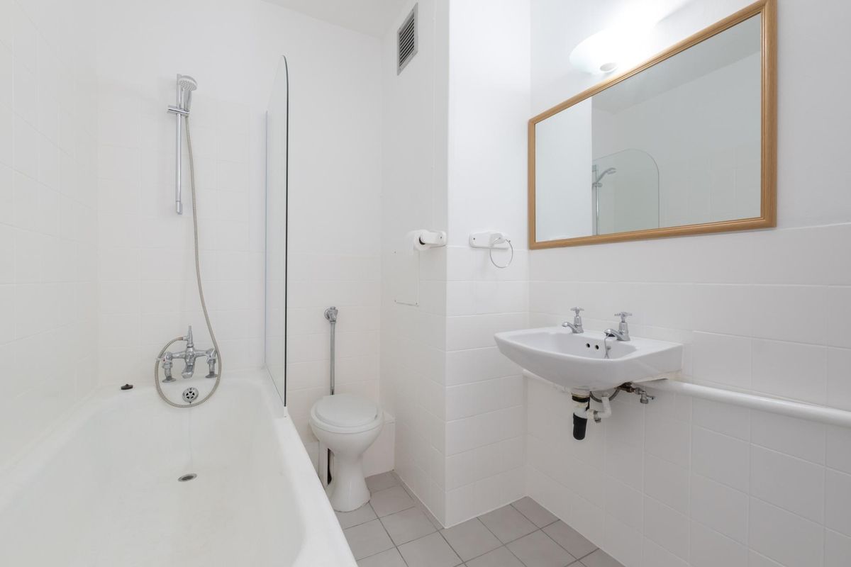 A clean, functional bathroom featuring a white bathtub with shower mixer, wall-mounted basin, and neutral tiled finishes throughout.