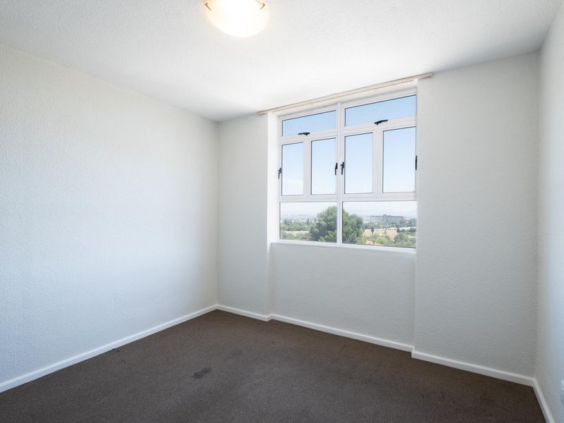 A bright, empty bedroom featuring neutral walls, carpeted flooring, and a large window with views of the surrounding area.