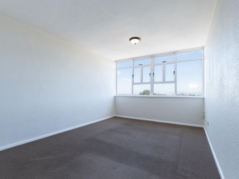 A bright, empty bedroom featuring neutral carpeted flooring, textured walls, and a large window providing natural light.