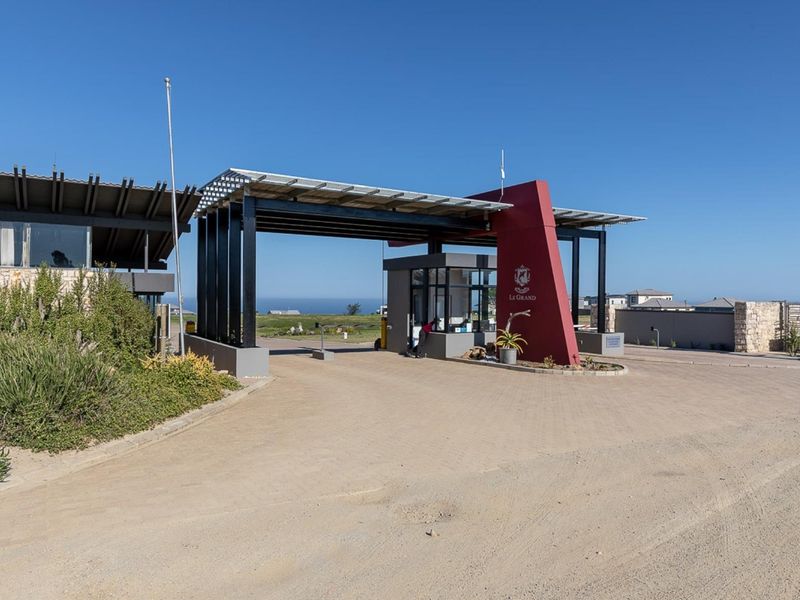 The entrance to Le Grand estate featuring a modern security gatehouse, paved driveway, and architectural steel structure with integrated solar panels.