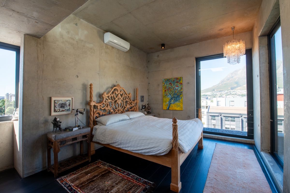 Modern bedroom featuring raw exposed concrete walls and ceiling, dark timber flooring, and expansive windows offering scenic mountain and urban views.