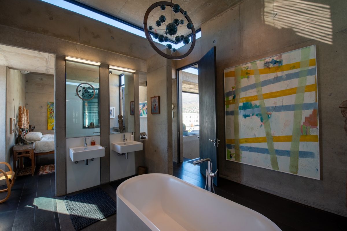 Modern industrial-style bathroom featuring exposed concrete finishes, a sleek freestanding bathtub, wall-mounted double vanity, and high-level clerestory windows for natural light.