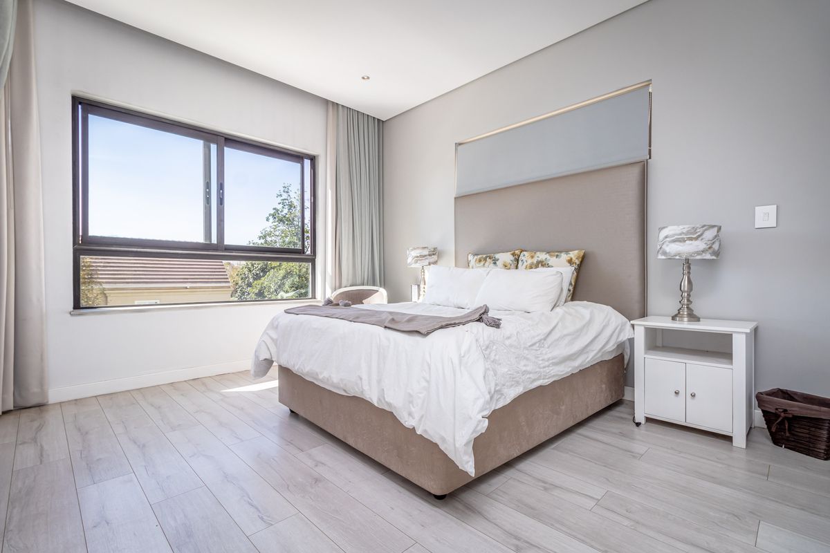 A bright and modern bedroom featuring light wood-look flooring, a large window with natural light, and a built-in headboard feature.