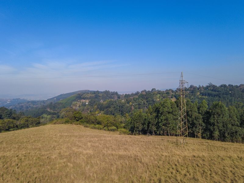 An aerial view of a vacant residential plot featuring rolling grassy terrain, mature trees, and an overhead electricity pylon.