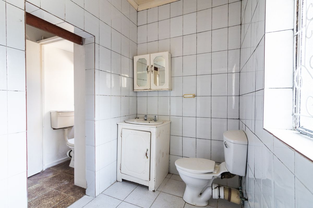 A dated bathroom featuring white wall tiles, a vanity basin, and a toilet, with an adjacent secondary toilet area visible through the doorway.