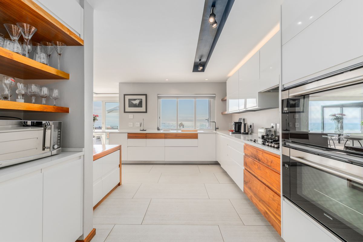 Modern kitchen featuring high-gloss white cabinetry, warm wood accents, integrated Siemens ovens, and sleek tiled flooring with a view toward the ocean.