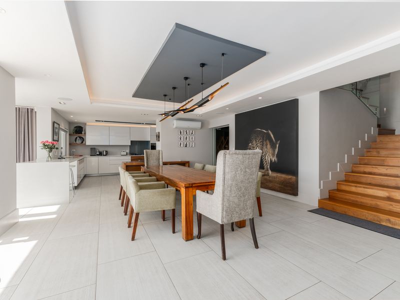 Modern open-plan living and dining area featuring a sleek kitchen, tiled flooring, recessed ceiling detail, and a timber staircase with glass balustrade.