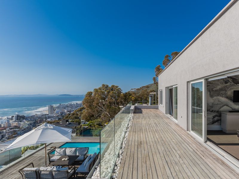 Expansive wooden deck balcony featuring a glass balustrade, overlooking a swimming pool with panoramic coastal and city views.