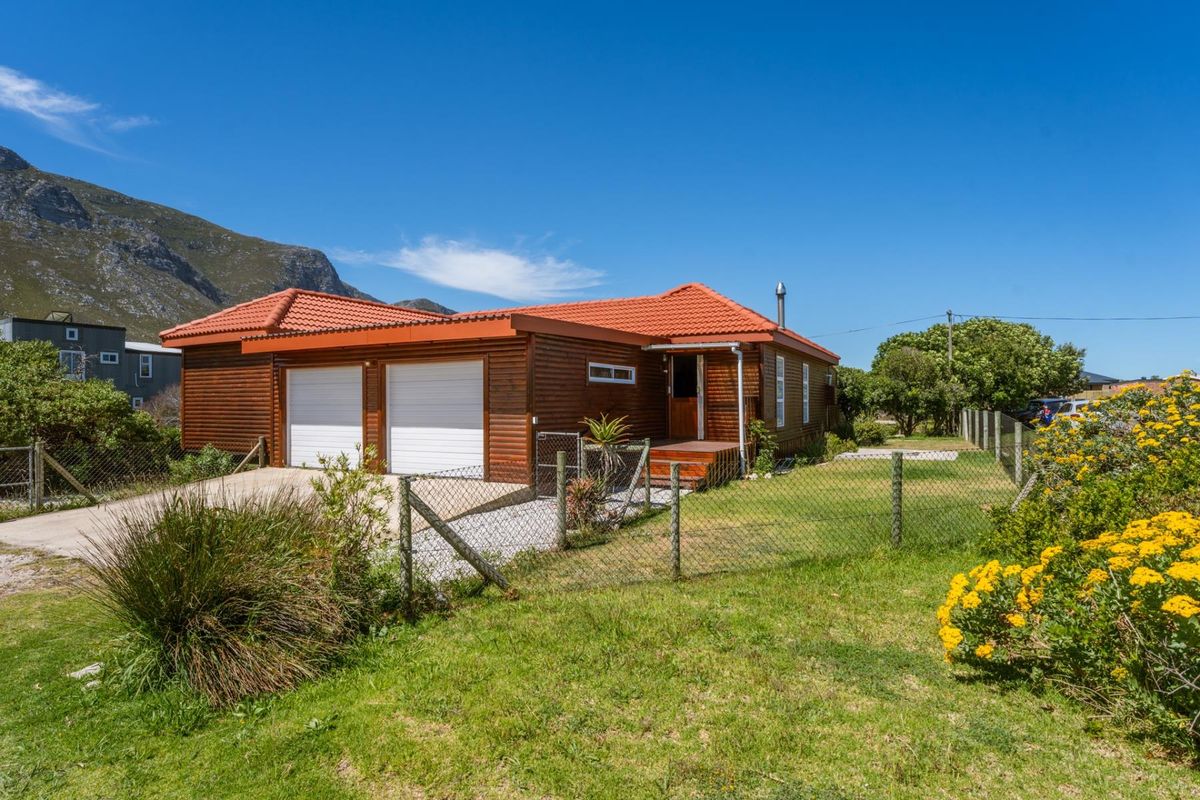 Front view of a timber-clad house featuring a double garage, tiled roof, and a small timber deck entrance, set within a fenced garden.