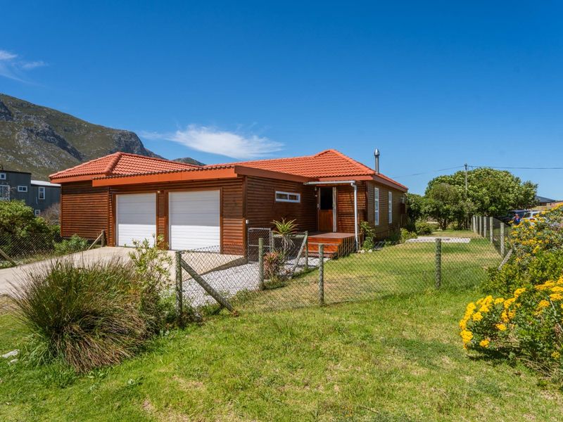 Front view of a timber-clad house featuring a double garage, tiled roof, and a small timber deck entrance, set within a fenced garden.