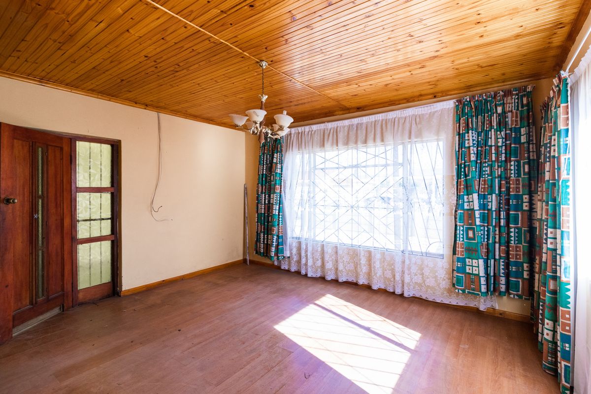 A spacious living room featuring a timber-paneled ceiling, large window with security bars, and wooden flooring, offering a blank canvas for renovation.