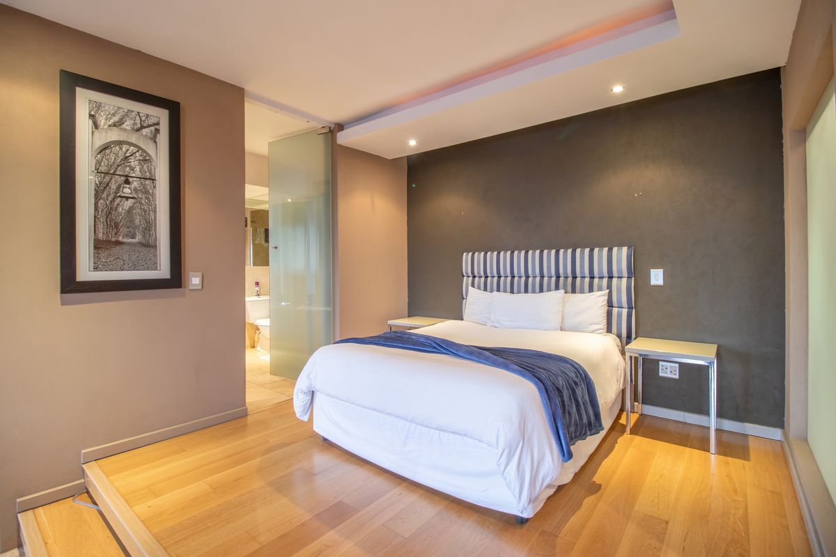 A modern bedroom featuring hardwood flooring, a recessed ceiling with integrated lighting, and an en-suite bathroom accessed via a glass door.