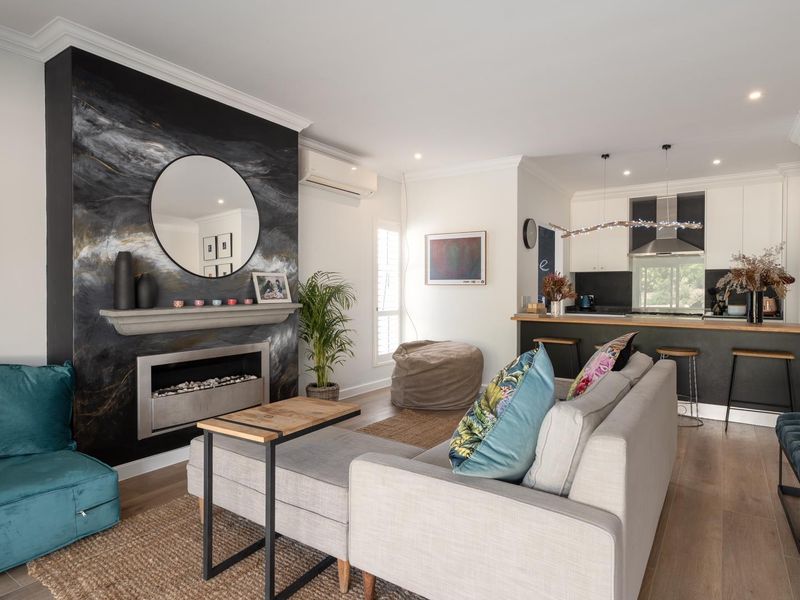 Open-plan living and kitchen area featuring a striking feature fireplace, modern kitchen island, timber flooring, and recessed lighting throughout.