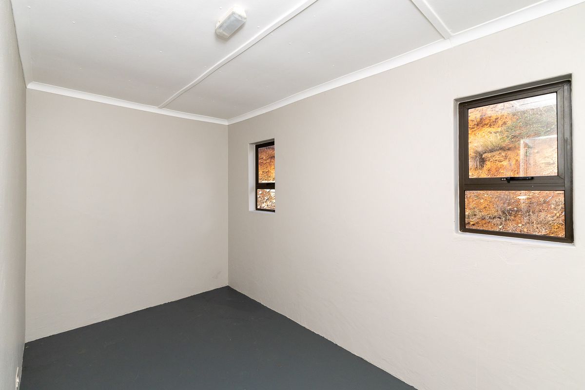 A clean, empty bedroom featuring neutral painted walls, dark painted flooring, and two aluminium-framed windows with views of the surrounding landscape.