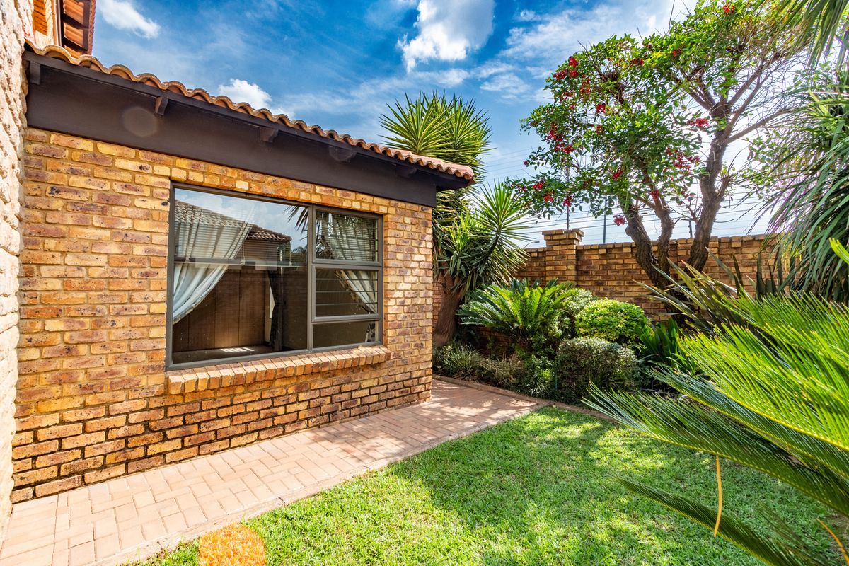 A well-maintained rear garden featuring a brick-paved walkway, established landscaping, and a face-brick exterior wall with a large window.
