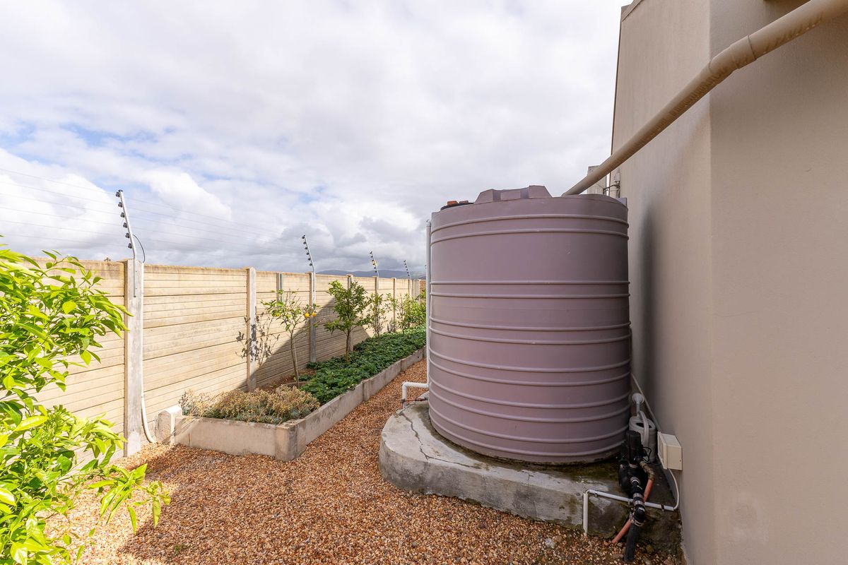 Side garden area featuring a large water storage tank on a concrete base, gravel ground cover, and a precast concrete boundary wall with electric fencing.