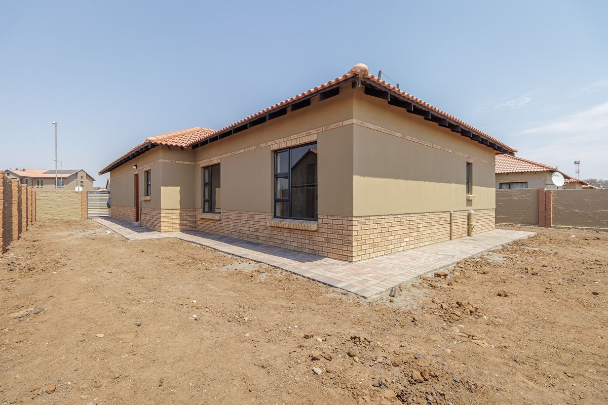 Exterior view of a single-story house featuring a tiled roof, brick and plaster facade, and paved perimeter walkway on an undeveloped plot.