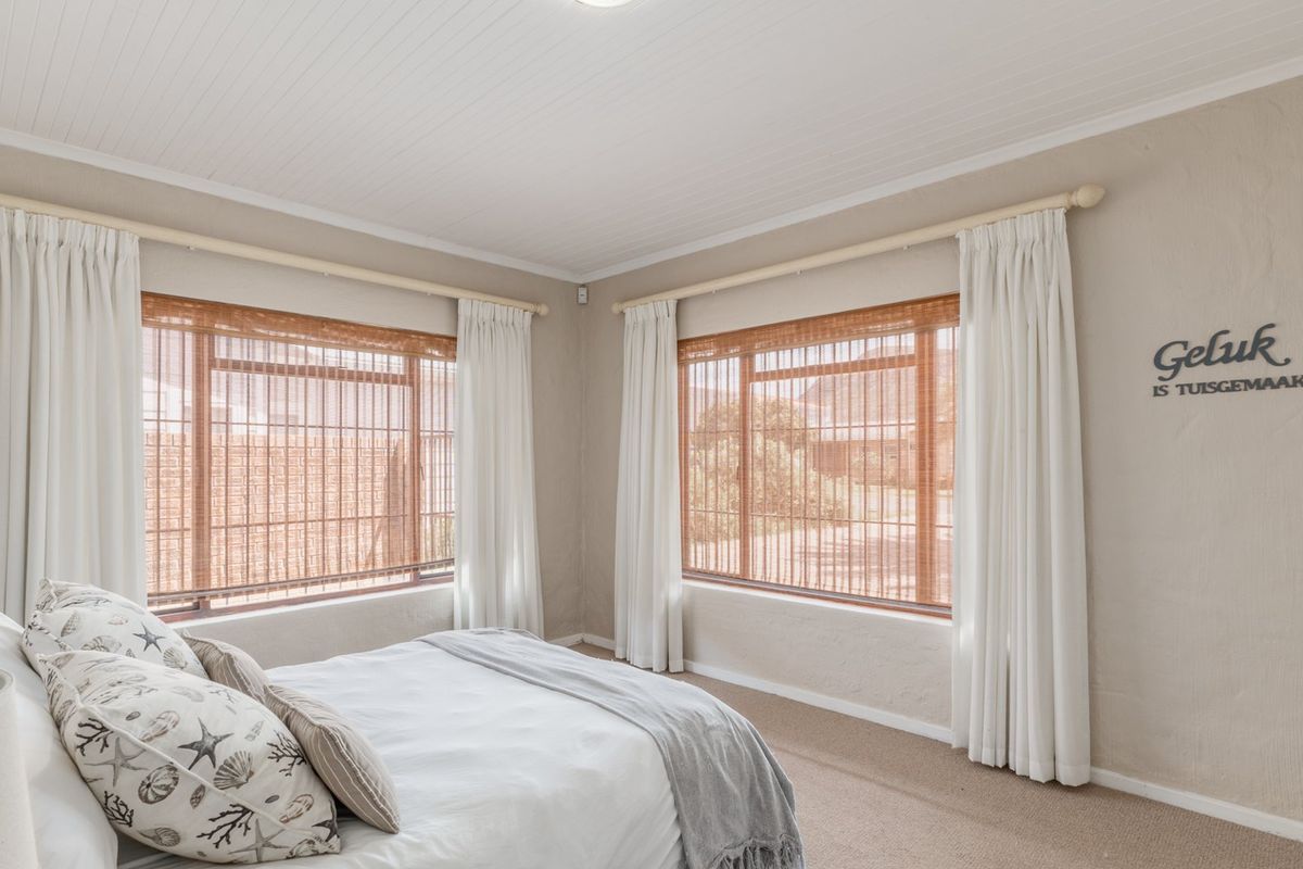 A bright bedroom featuring neutral wall tones, carpeted flooring, and large windows with fitted curtain rods and blinds.