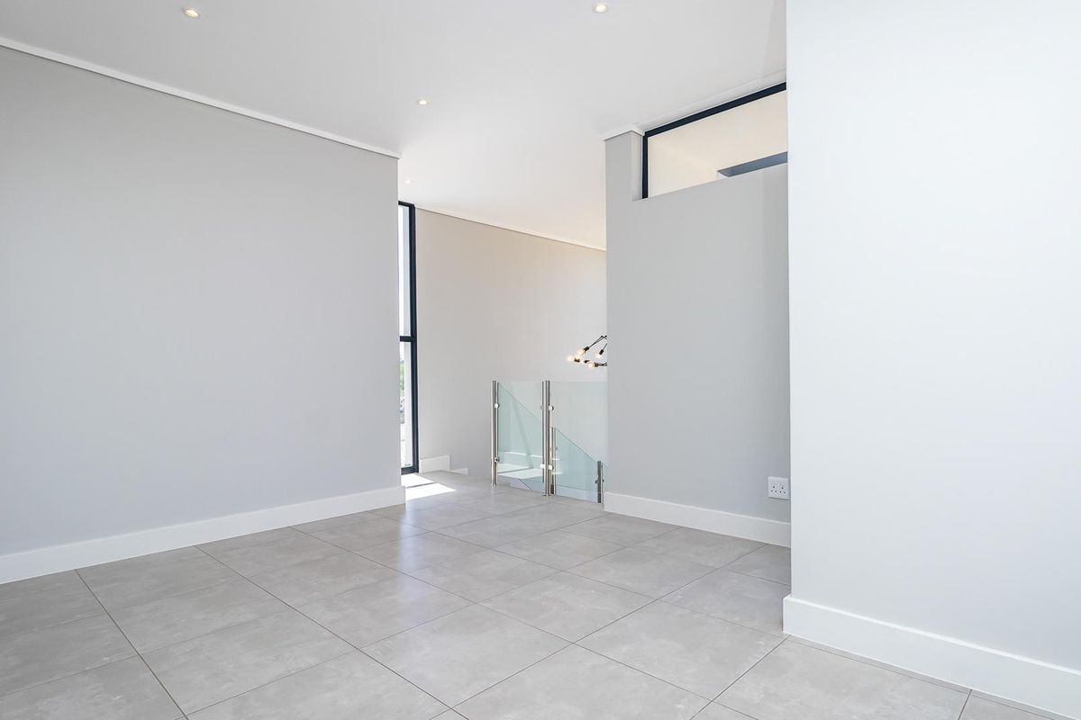 A bright, modern, and empty living space featuring neutral grey floor tiles, recessed ceiling lighting, and a contemporary glass balustrade staircase.
