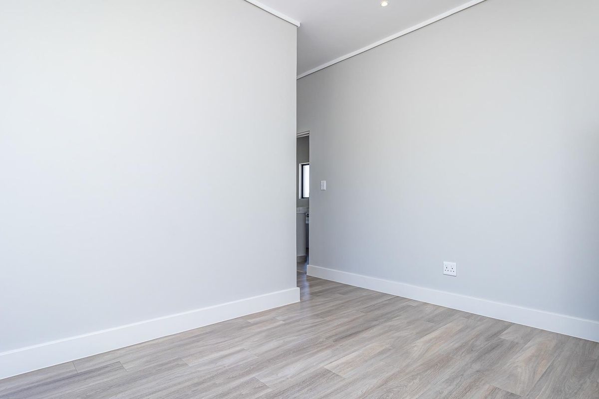 A bright, empty bedroom featuring modern light grey walls, white skirting boards, and wood-look laminate flooring with a doorway leading to an adjacent space.