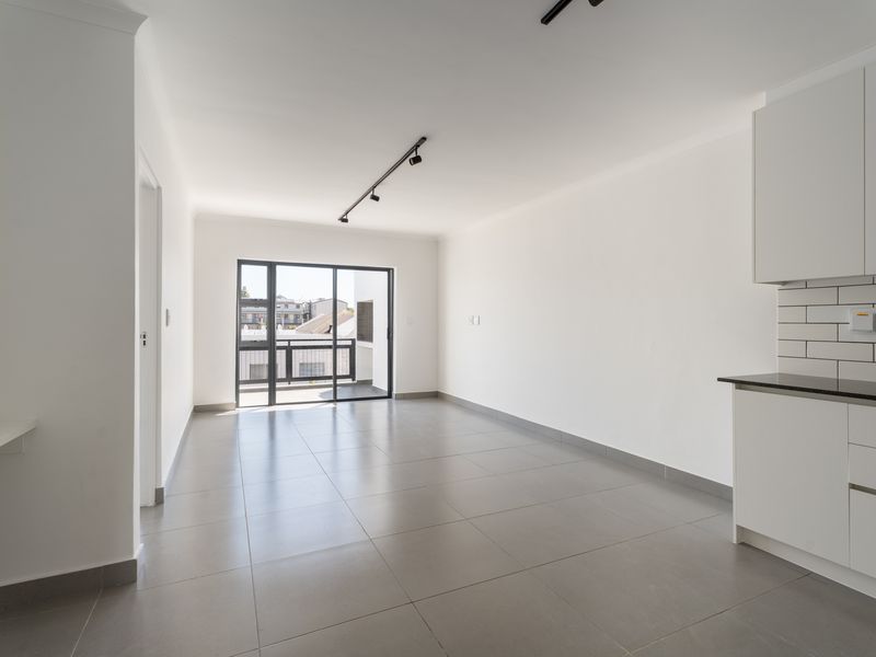A bright, modern open-plan living and kitchen area featuring neutral tiled floors, contemporary track lighting, and direct access to a private balcony.
