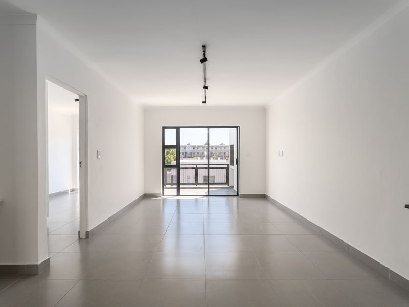 A bright, empty open-plan living area featuring neutral tiled flooring, modern track lighting, and a sliding glass door leading to an outdoor balcony.